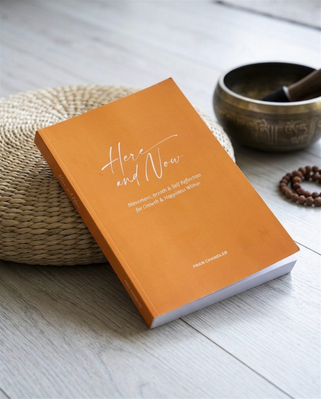 Here and Now leaning against a woven pouf beside a singing bowl and mala beads